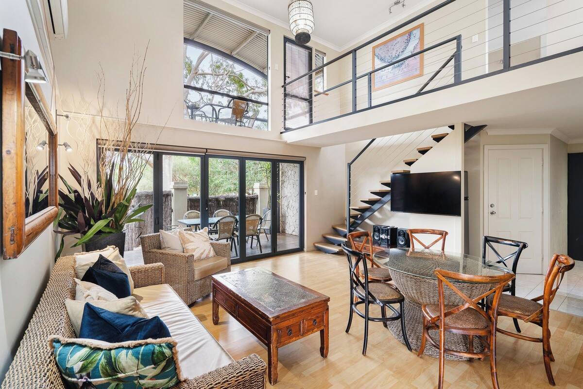 A spacious living area with a sofa, coffee table, and dining set. A staircase leads to an upper level with a balcony.