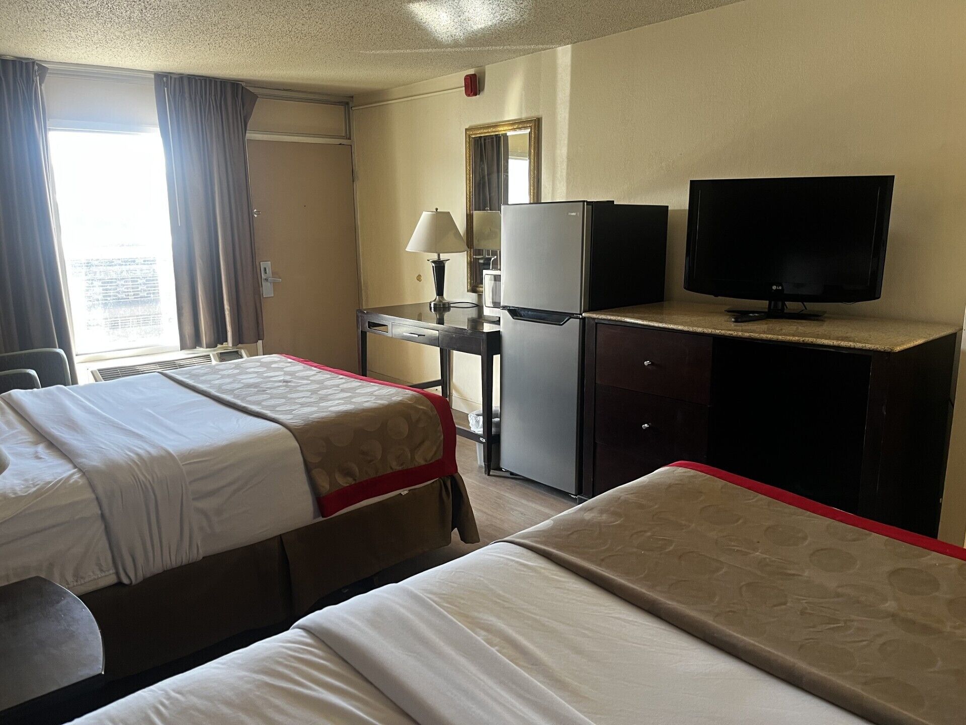 A hotel room with two beds, a desk with a refrigerator, a television, and a window with curtains.