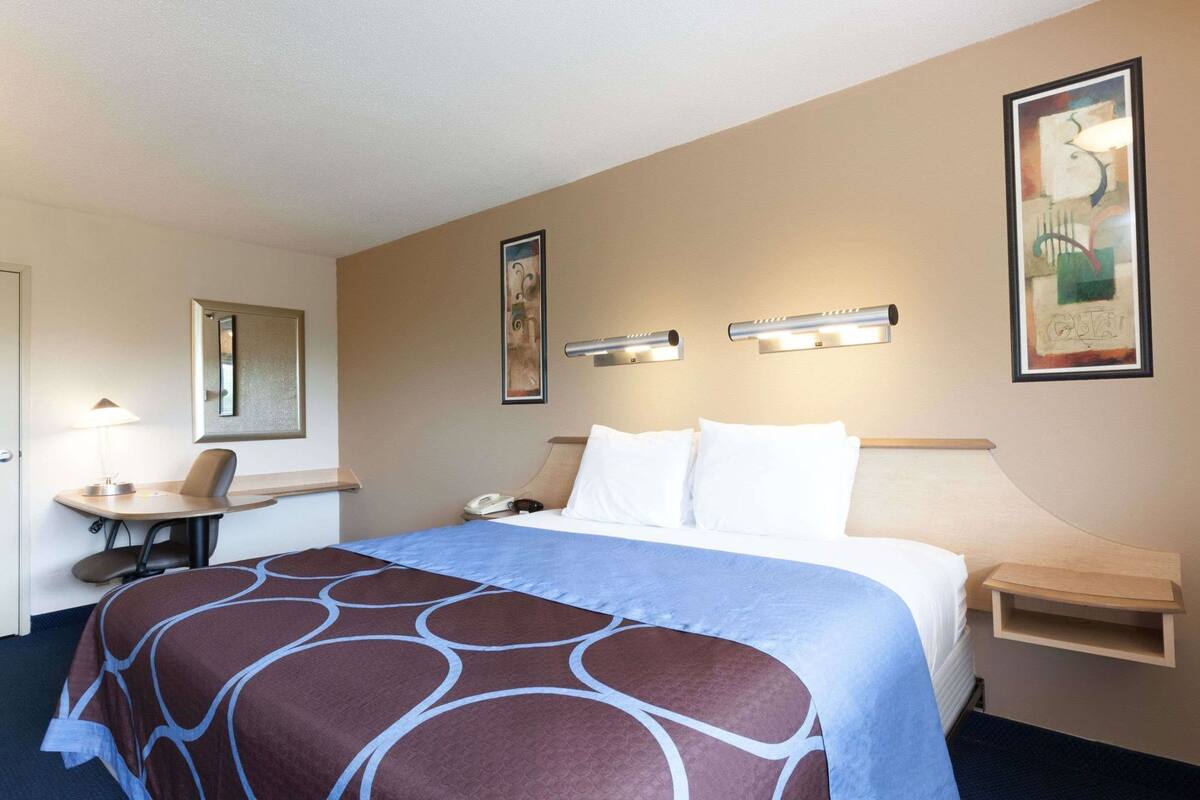 A hotel room with a bed, desk, chair, bedside table, lamp, and artwork on the walls.