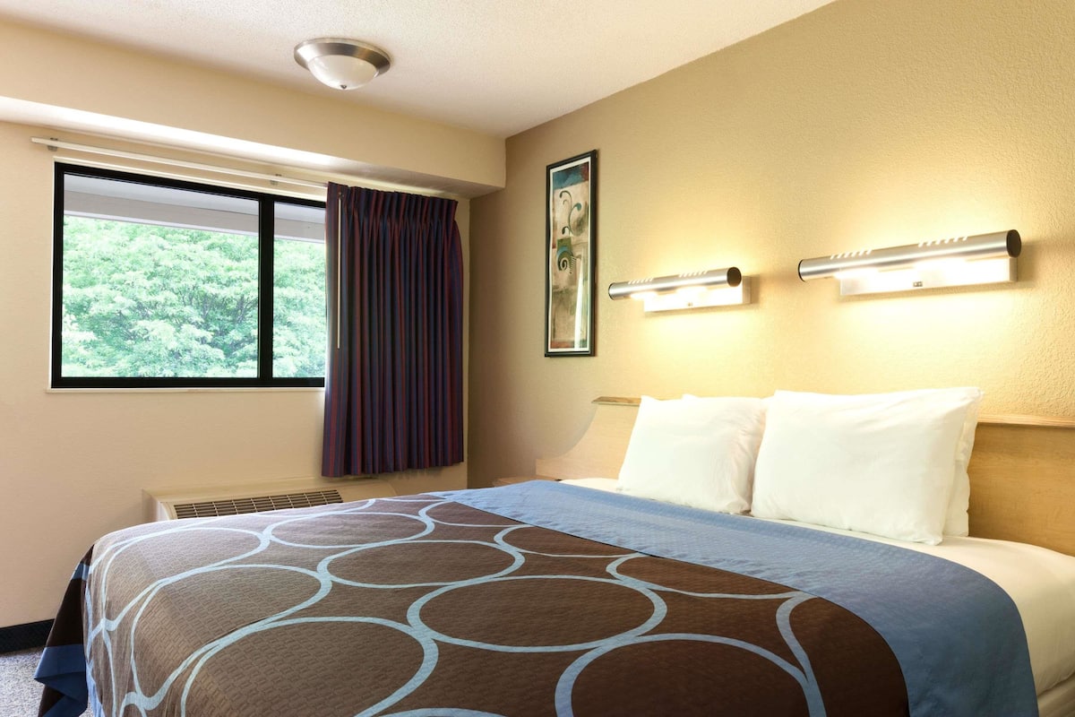 A hotel room with a large bed, a window with curtains, a wall-mounted picture, and two wall sconces.