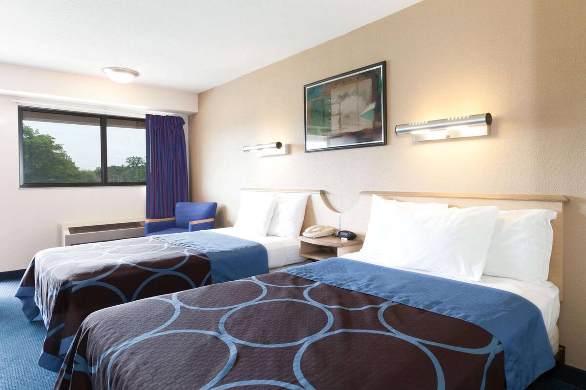 A hotel room with two beds, a window with curtains, a painting on the wall, and a bedside lamp.