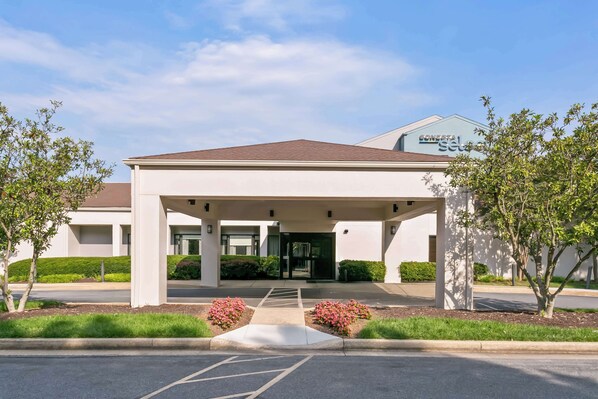 A modern hotel entrance with a covered walkway, a parking lot, and landscaped areas with shrubs and flowers.