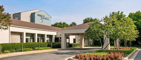 A modern hotel entrance with a covered walkway, landscaped gardens, and a clear sky.