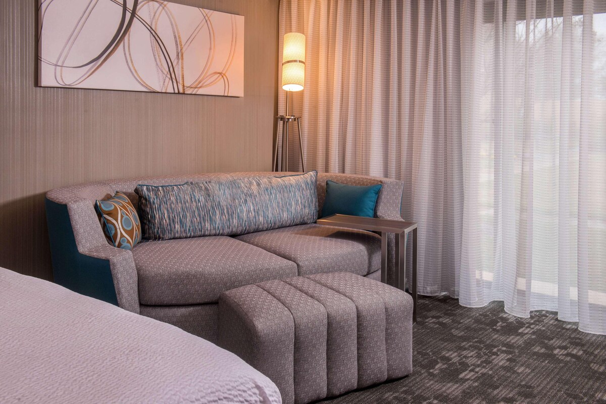 A modern hotel room with a sectional sofa, a floor lamp, and sheer curtains.