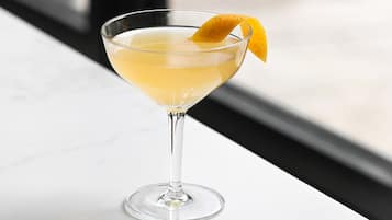 Een cocktail in een martini-glas, versierd met een citroenschil, op een witte marmeren oppervlak.