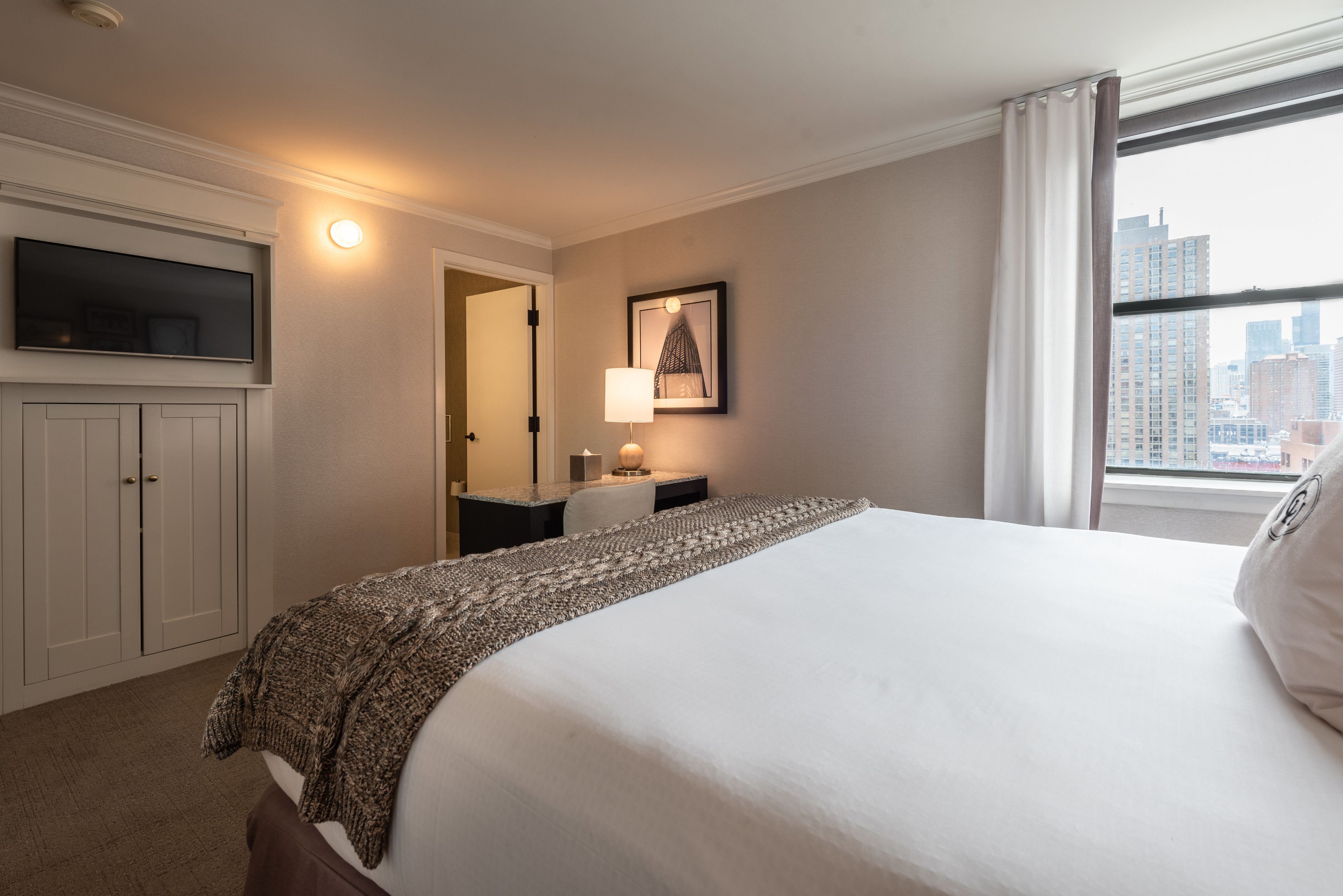 Una habitación de hotel con cama, televisor montado en la pared, mesita de noche con lámpara y ventana con vista a la ciudad.