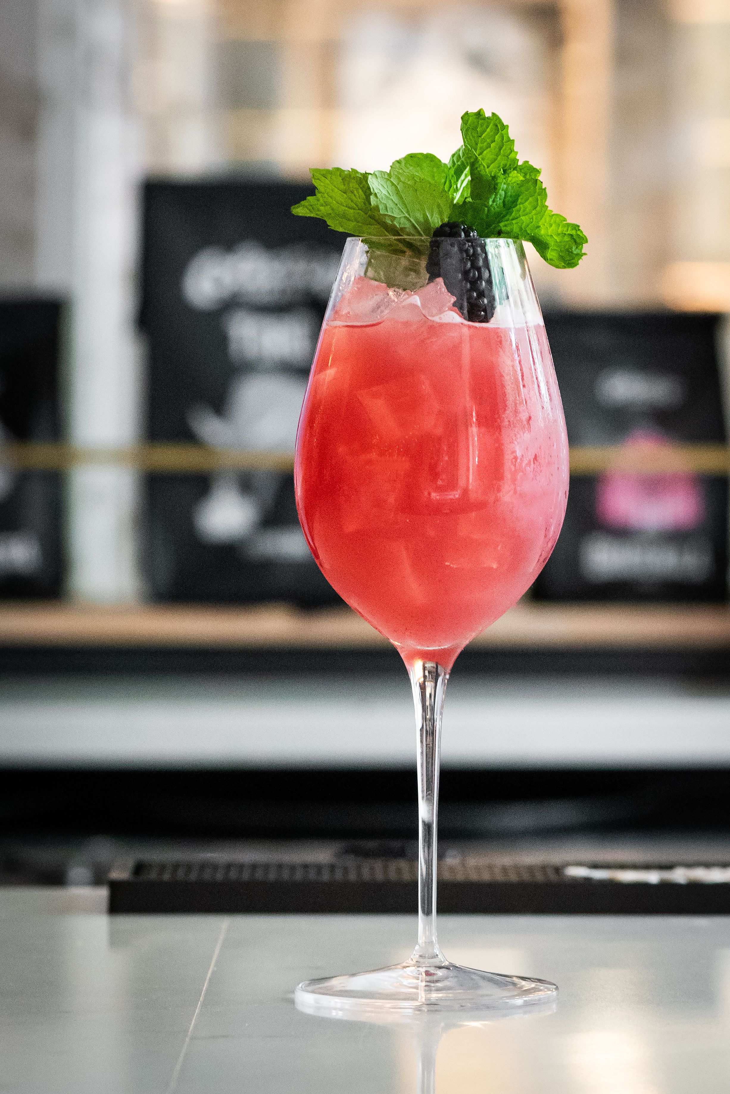 A cocktail in a stemmed glass with a mint garnish and a blackberry, placed on a bar counter with a blurred background.