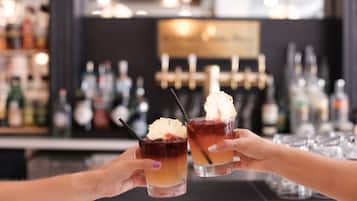 Two hands holding cocktail glasses with ice cream on top, a bar with various bottles in the background.