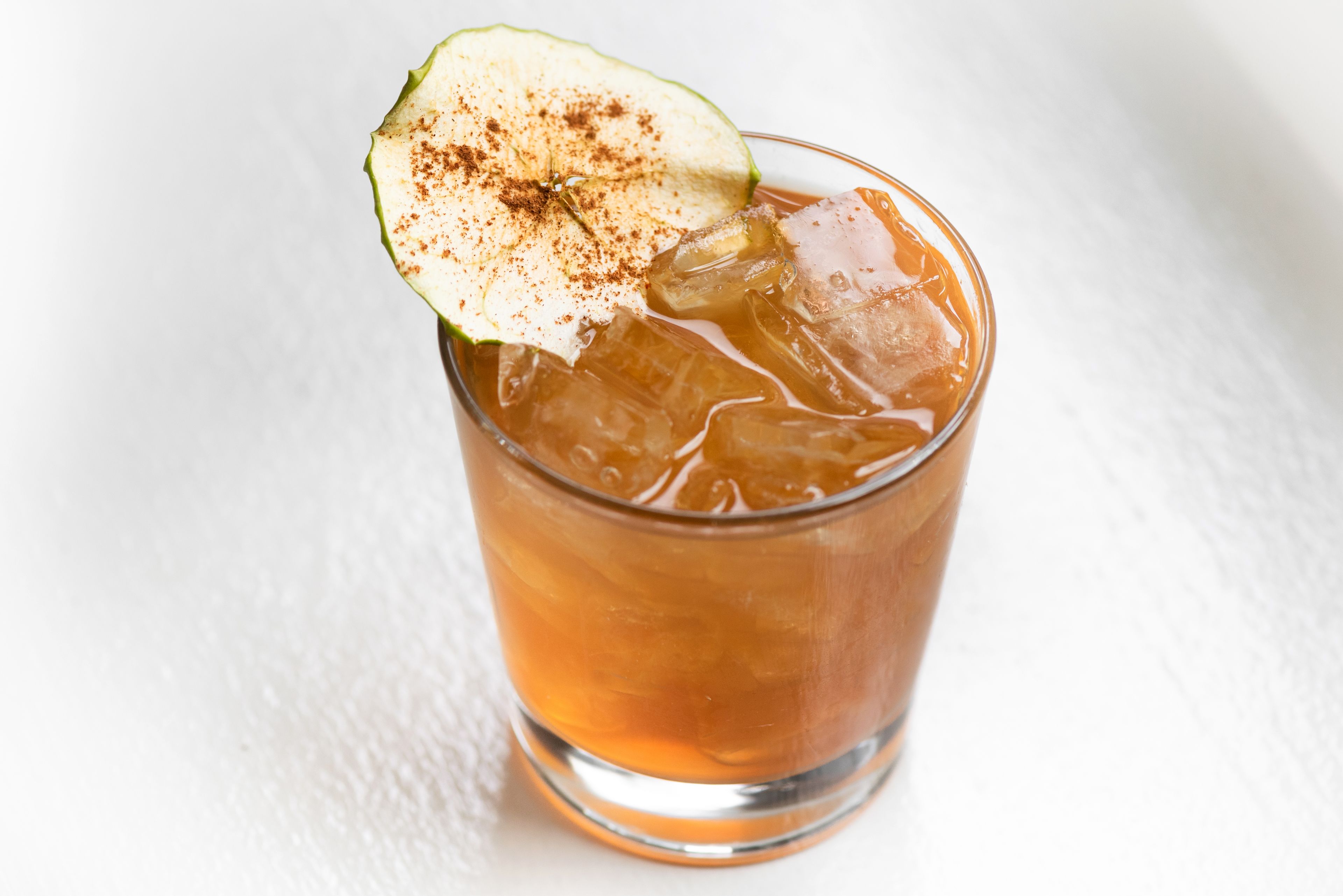 Un verre d’une boisson ambrée, avec des glaçons et une tranche de pomme saupoudrée de cannelle sur le bord.
