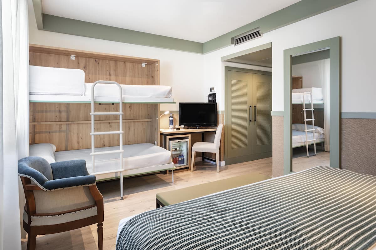 Quarto de hotel compacto com beliches, uma escrivaninha com computador, uma cadeira e uma cama.