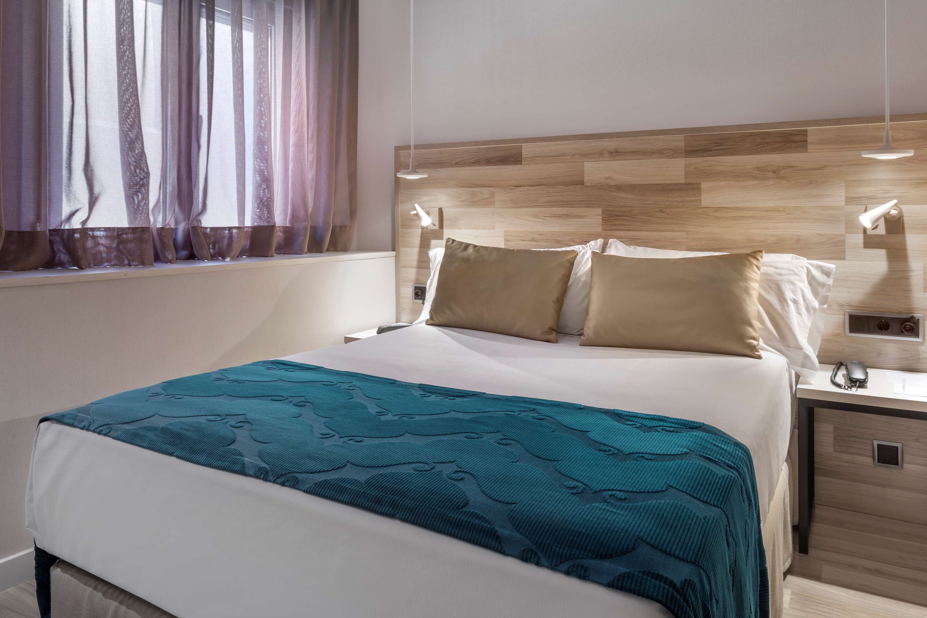 Una cama bien hecha con una colcha estampada, cabecero de madera, lámparas de noche y una mesita de noche con un teléfono y cargador.