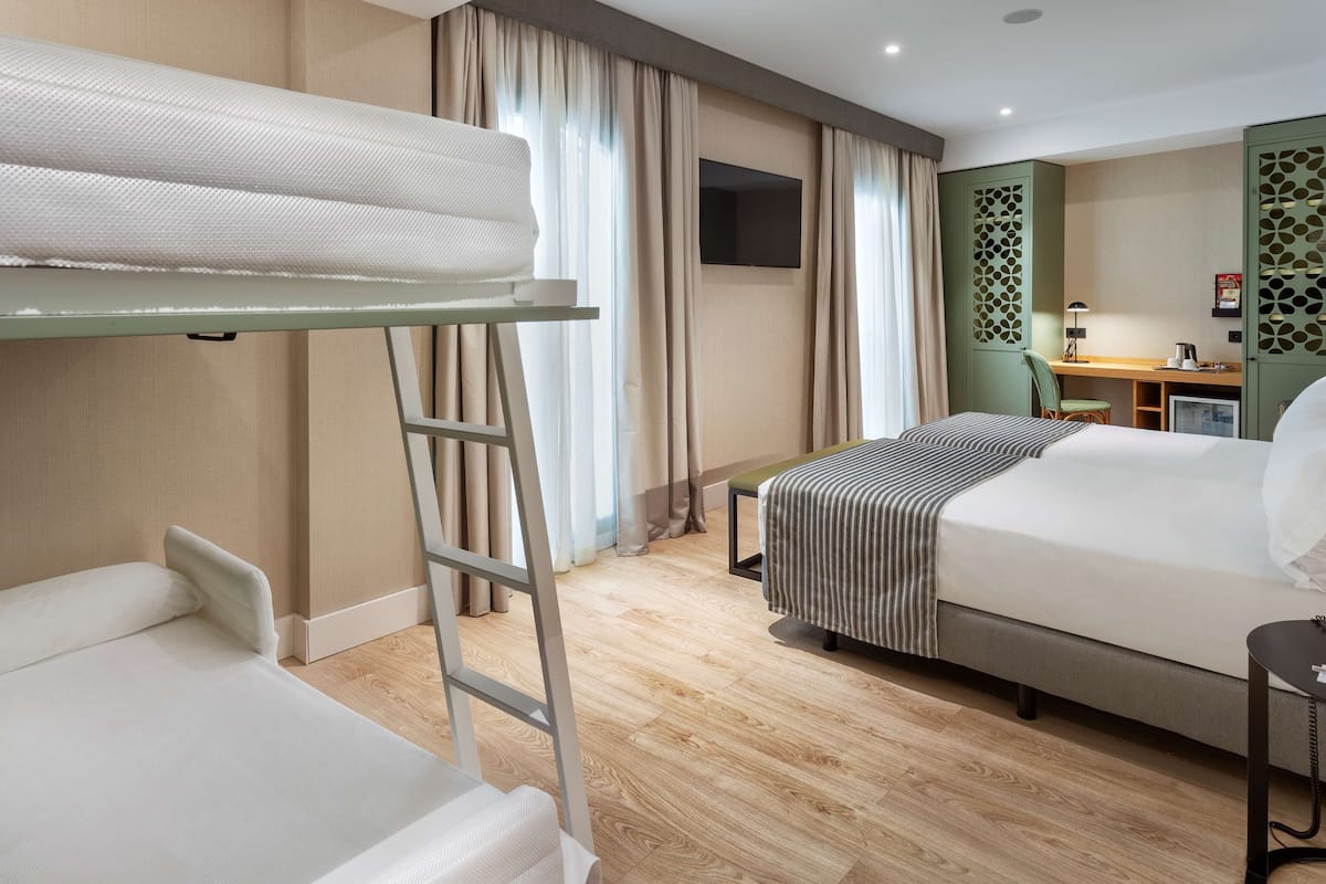Quarto de hotel com uma cama beliche, uma mesa com cadeira, uma televisão e uma pequena mesa com abajur.
