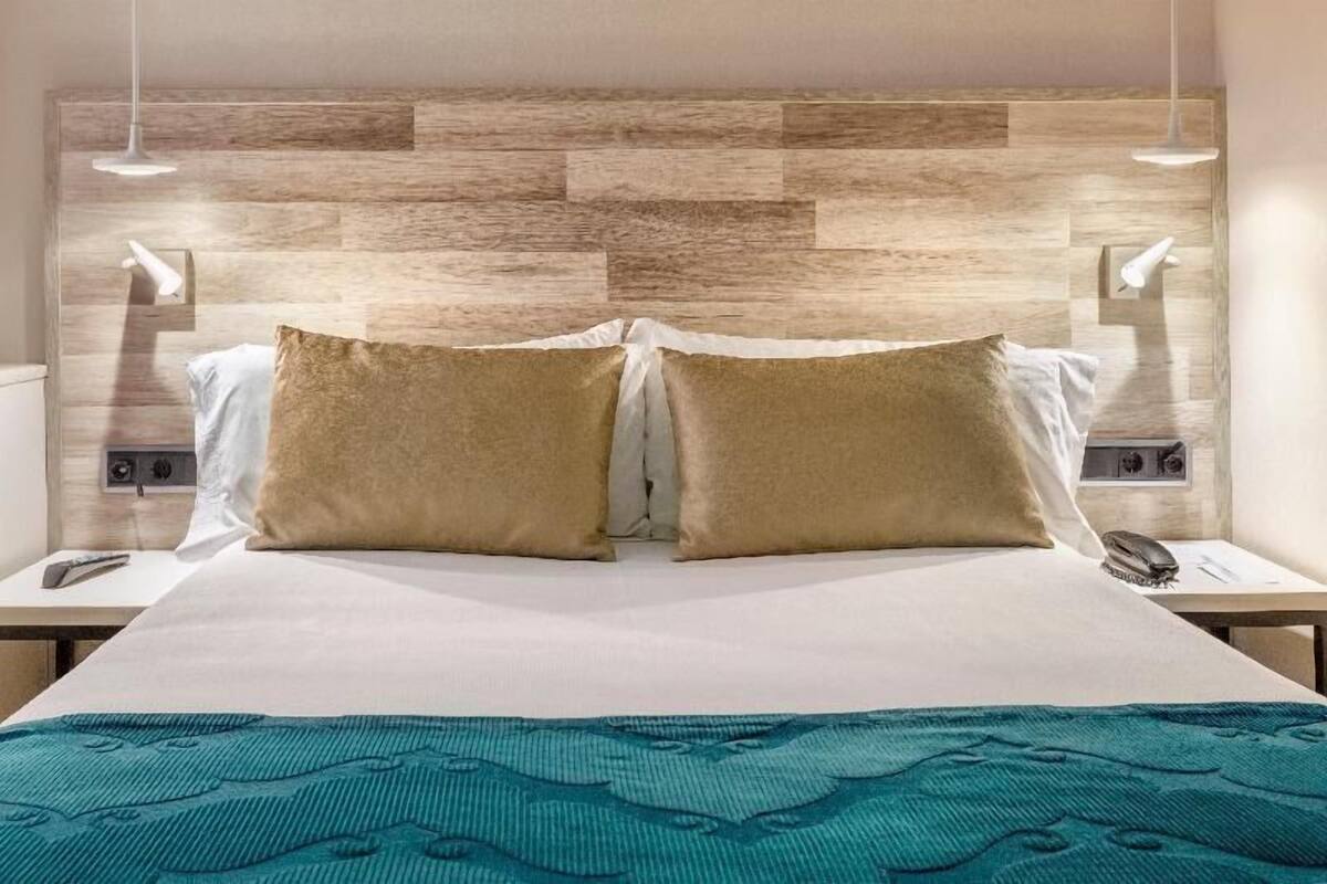 Quarto de hotel com cama, duas luminárias de apoio, telefone e luminária fixada na parede.