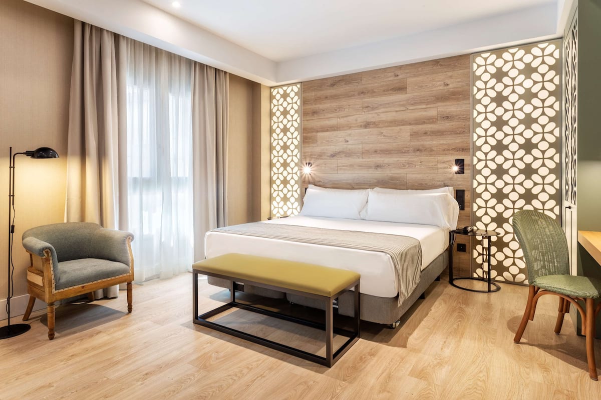 Quarto de hotel moderno com uma cama grande, um banco, duas cadeiras e um painel decorativo na parede.