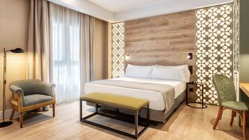 A modern hotel room with a large bed, a bench, two chairs, and a decorative wall panel.