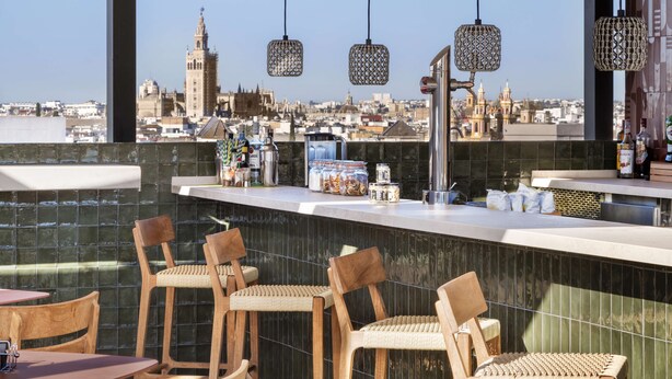 Un bar en la azotea con vistas a la ciudad, sillas de mimbre y una barra con botellas y vasos.