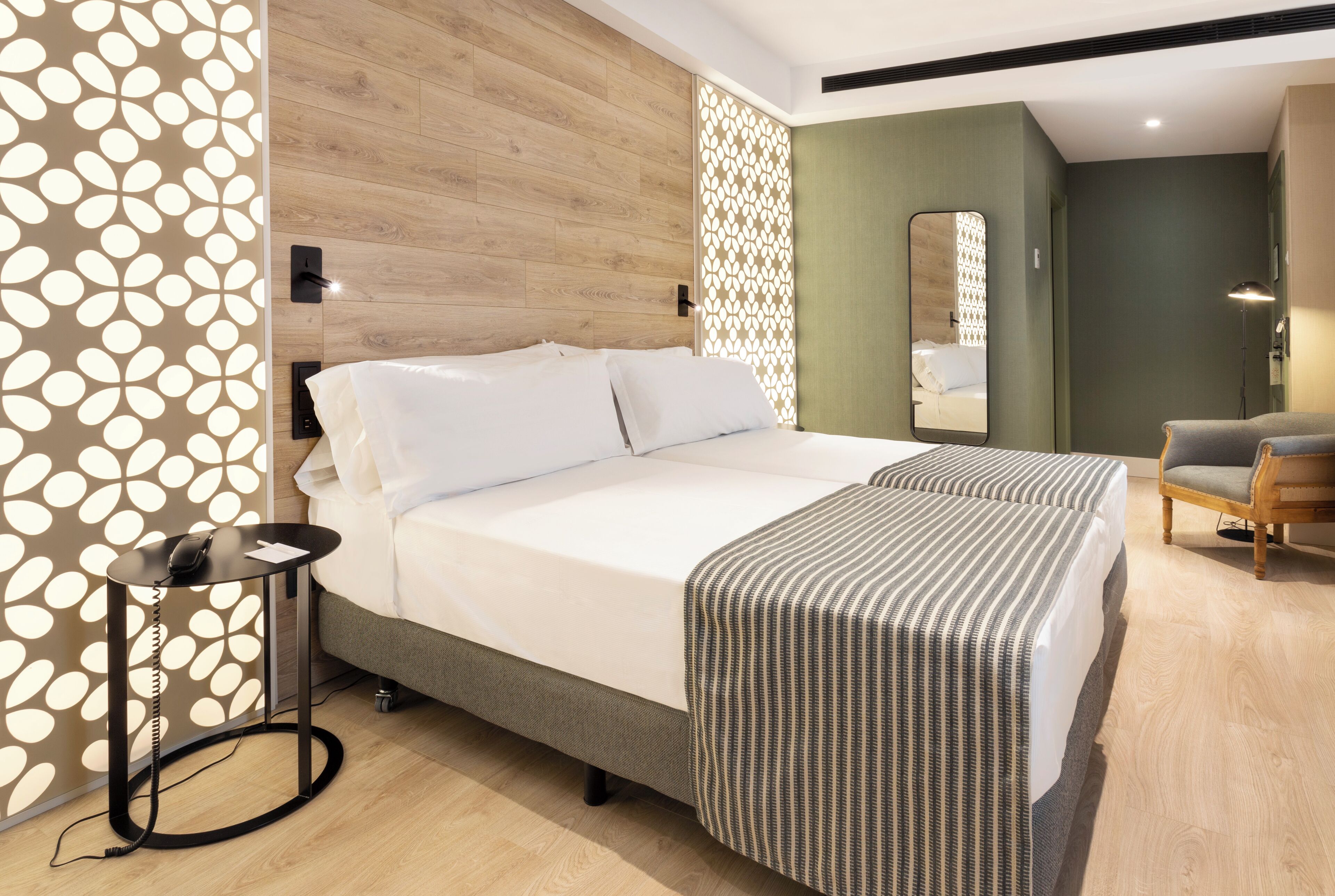 Ein modernes Hotelzimmer mit einem hölzernen Kopfteil, einem großen Bett mit weißen Bettwäsche, einer gestreiften Tagesdecke, einem kleinen Nachttisch mit einem Telefon und einem Stuhl.