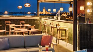 Un bar en la azotea con vistas a una torre histórica, muebles de madera y luces colgantes.