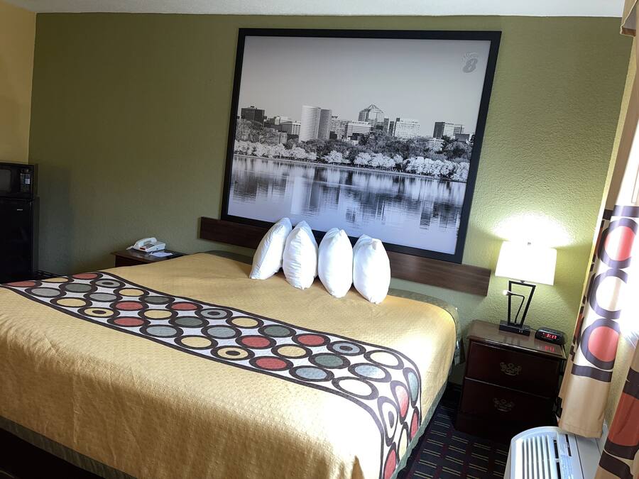 Una habitación de hotel con cama, mesita de noche, lámpara, reloj y una fotografía enmarcada de un paisaje urbano en la pared.