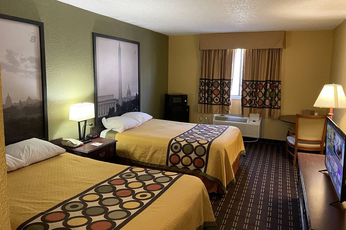 Ein Hotelzimmer mit zwei Betten, einem Fernseher, einem Schreibtisch, einem Stuhl und einem fensterbepflanzten Fenster.
