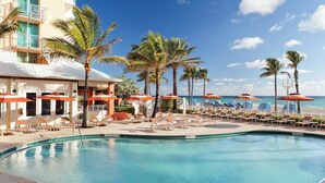 Zona de piscina con sillones de playa y sombrillas, palmeras y un edificio de fachada blanca.