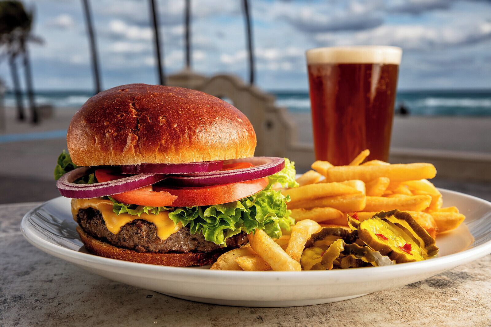 En tallrik med en hamburgare, pommes frites och ett glas öl står på ett bord med utsikt över stranden i bakgrunden.