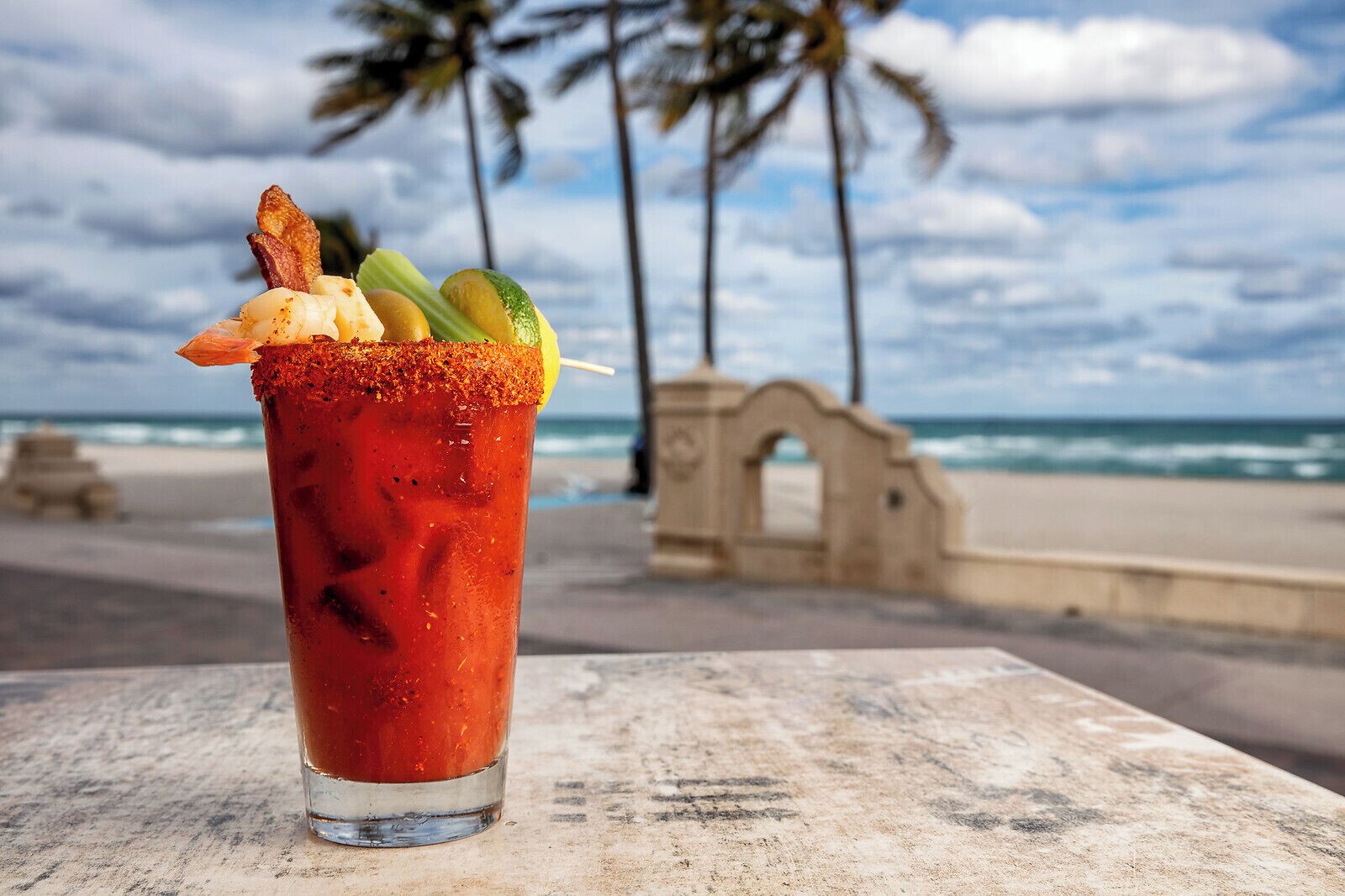 Un cóctel Bloody Mary adornado con tocino, apio y una cuña de limón, sobre una mesa con una playa y palmeras al fondo.