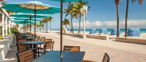 Een terras aan het strand met tafels en stoelen, parasols en palmbomen.
