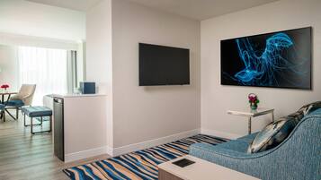 A modern hotel room with a flat-screen TV, a sofa, a dining area with a table and chairs, and a kitchen area with a dishwasher.