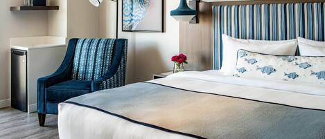 A modern hotel room with a large bed, a blue armchair, a bedside table with a vase of flowers, and a wall-mounted lamp.