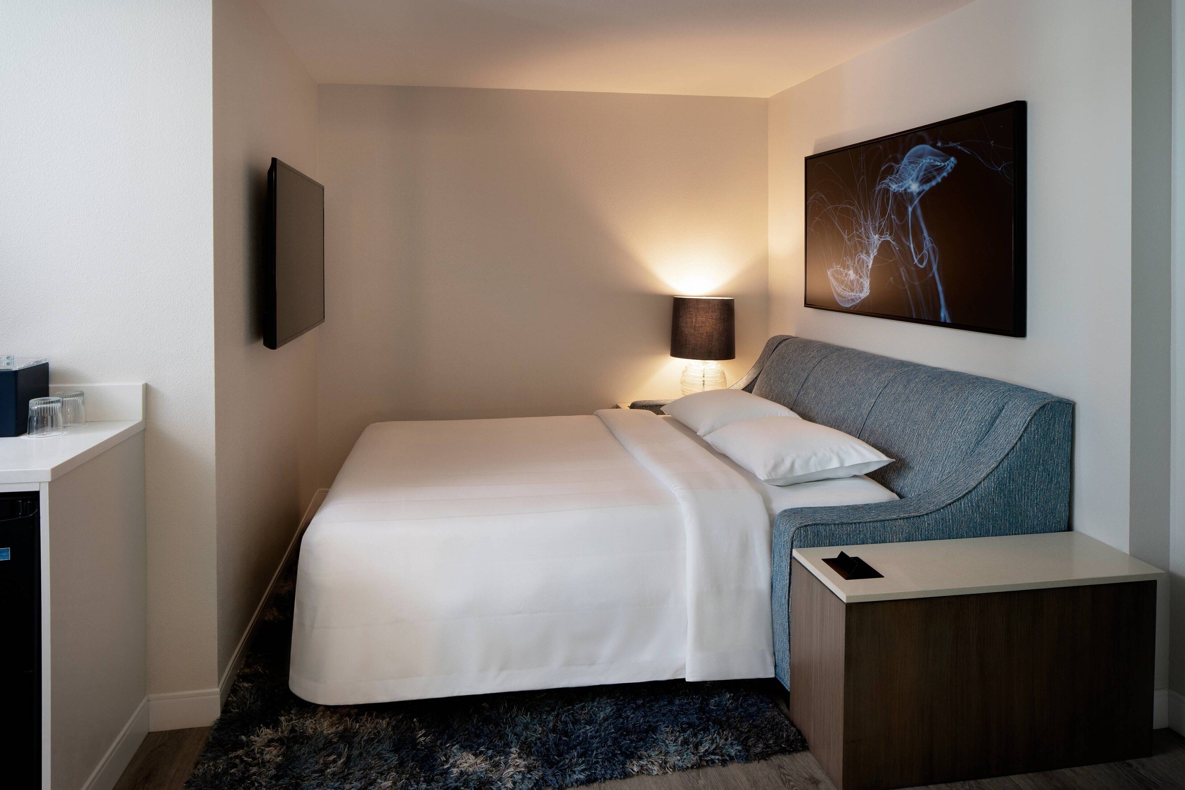 Una habitación de hotel moderna con una cama bien hecha, una mesita de noche, una lámpara y una obra de arte colgada en la pared.