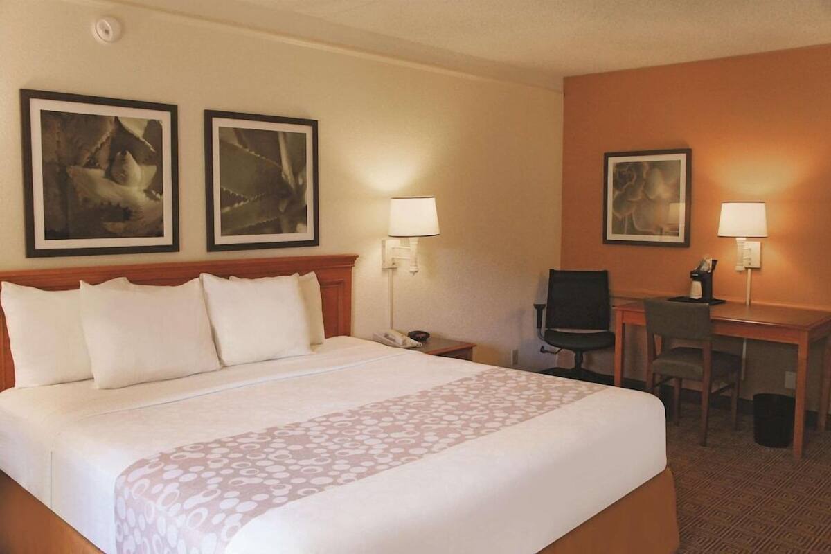 A hotel room with a bed, two wall-mounted lamps, a desk with a chair, and two framed pictures on the wall.