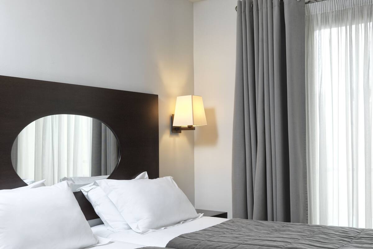 Habitación de hotel con cama, espejo redondo, lámpara de pared y ventana con cortinas.