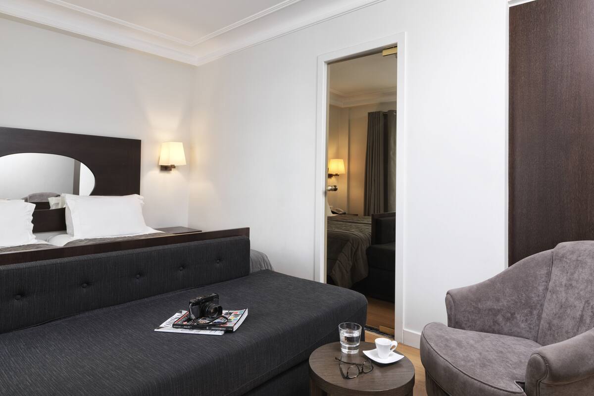 Una habitación de hotel moderna con una cama grande, una mesita redonda con una taza y vasos, una silla y una puerta que da a un baño.