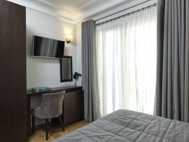A hotel room with a bed, a desk with a chair, a flat-screen TV, a mirror, and a window with curtains.