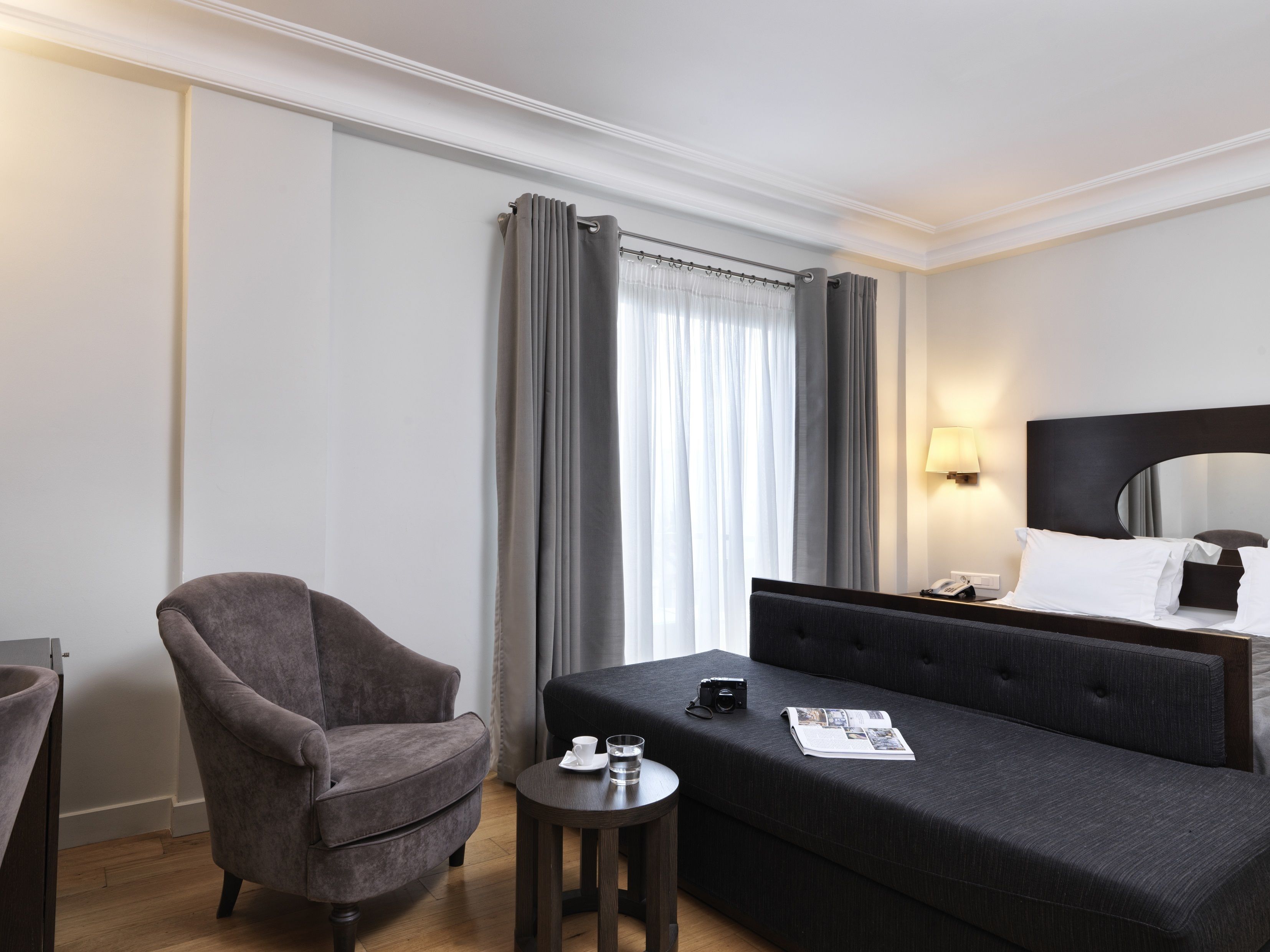 Een hotelkamer met een bed, een grijze fauteuil, een klein tafeltje met een kopje en een camera, en een raam met grijze gordijnen.
