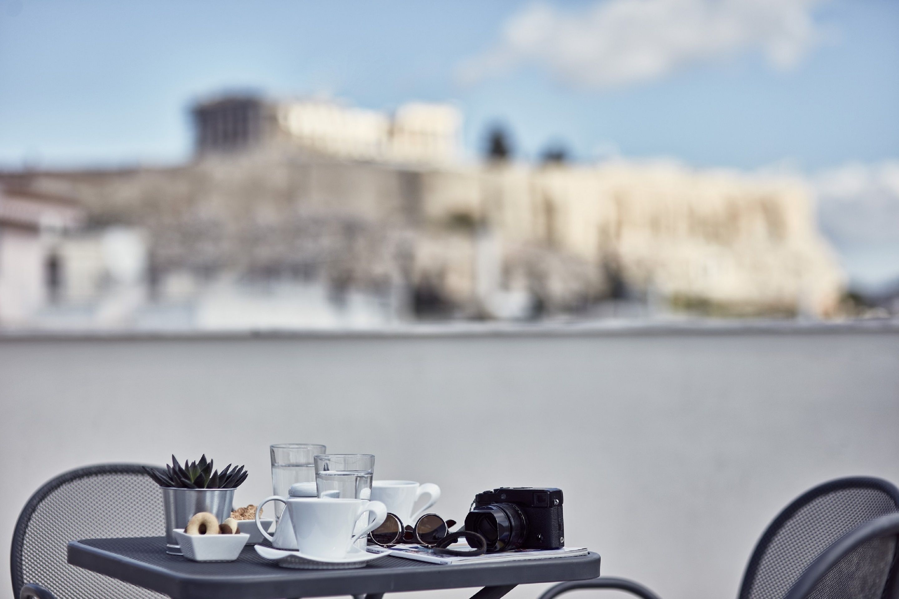 Una mesa con una cámara, tazas y una planta pequeña en una terraza con vista a un edificio histórico al fondo.