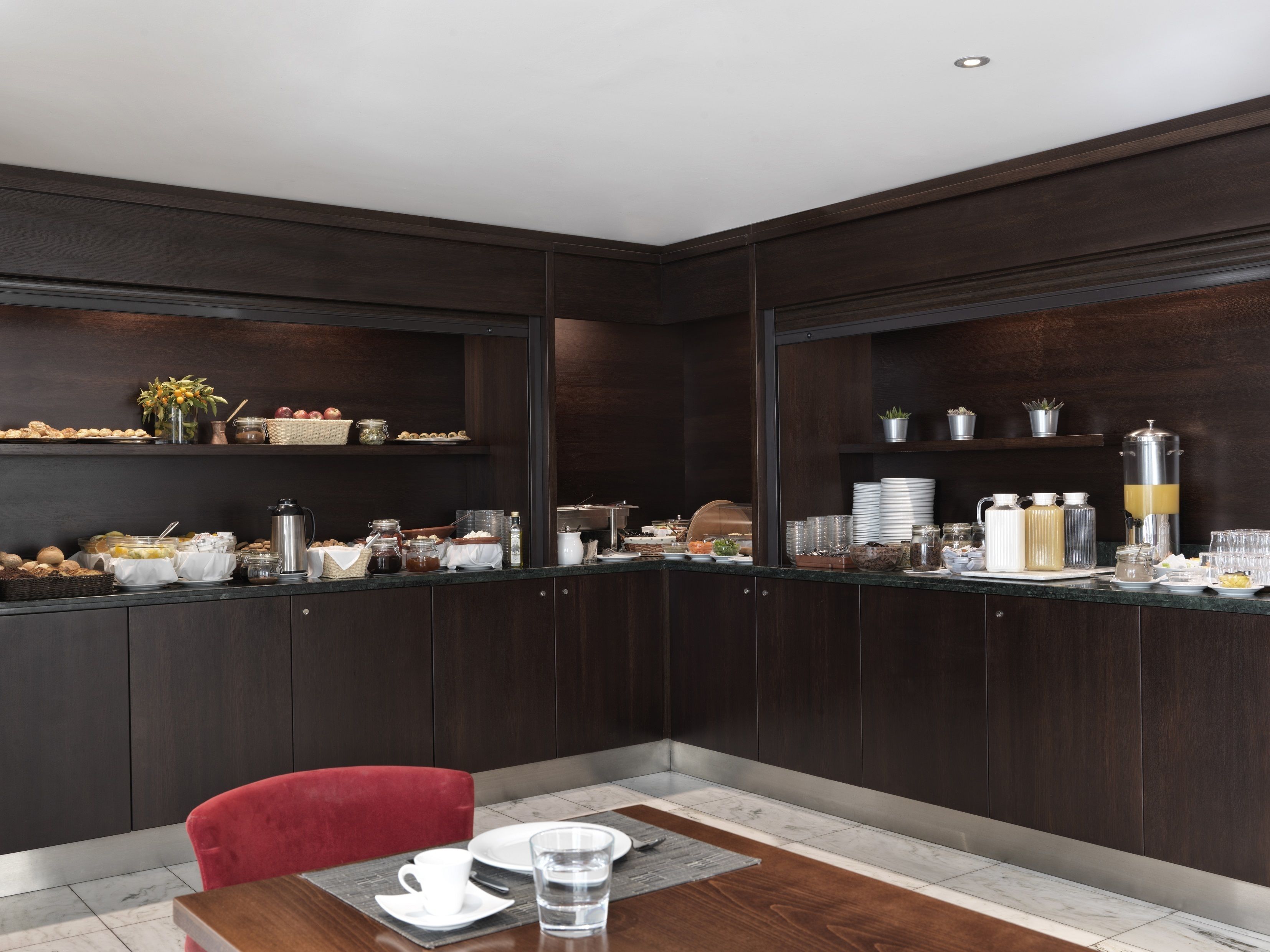 A modern kitchen with dark wood cabinets, a marble countertop, and a dining area with a red chair and a wooden table.