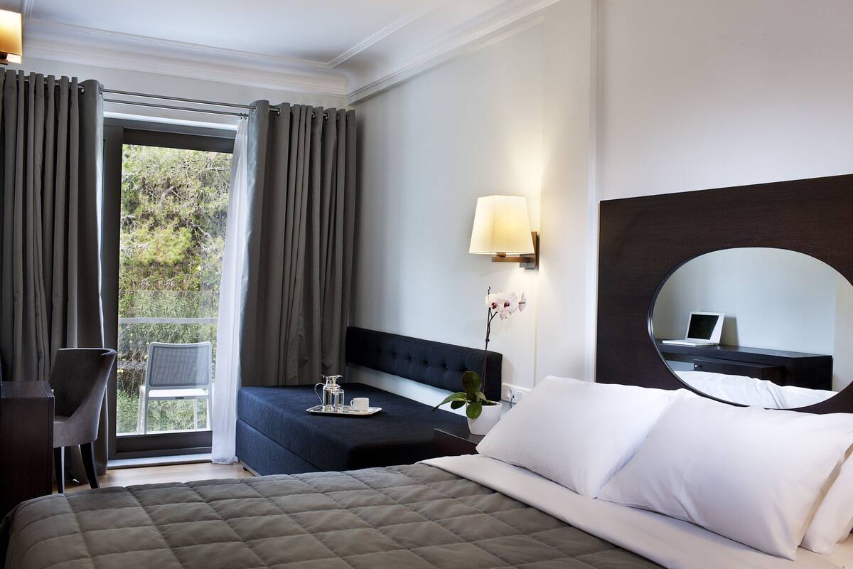 Habitación de hotel con cama, escritorio con computadora portátil, una silla, ventana con cortinas y una planta en maceta.