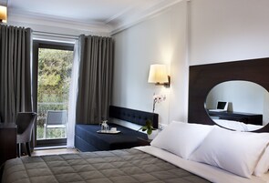 Quarto de hotel com uma cama, uma escrivaninha com um laptop, uma cadeira, uma janela com cortinas e uma planta em vaso.