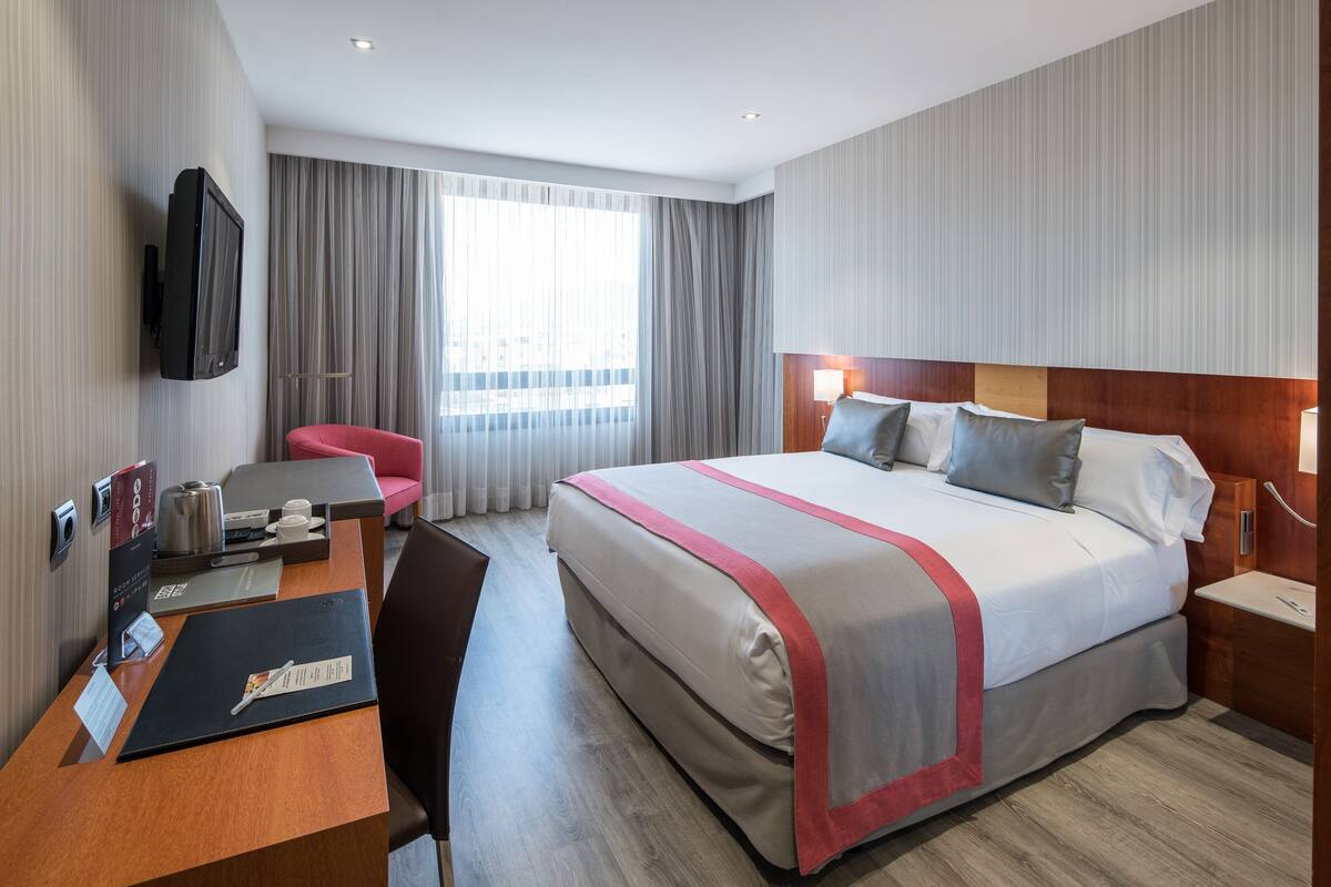 A hotel room with a bed, a desk, a chair, a TV, and a window with curtains.