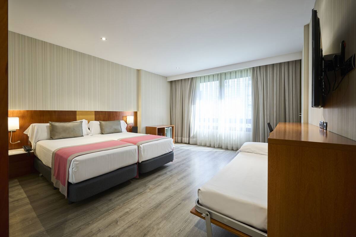 A hotel room with two beds, a desk, a TV, and a large window with curtains.