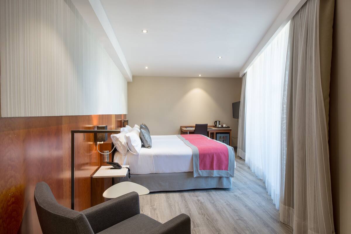 A hotel room with a bed, a desk, a chair, a small table, and a TV.