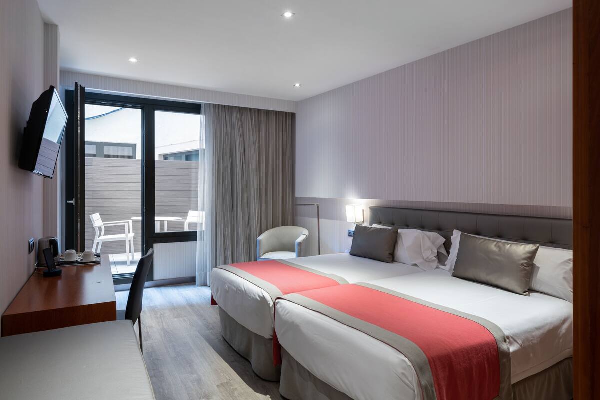 A hotel room with a large bed, a desk with a chair, a television mounted on the wall, and a sliding glass door leading to an outdoor area.