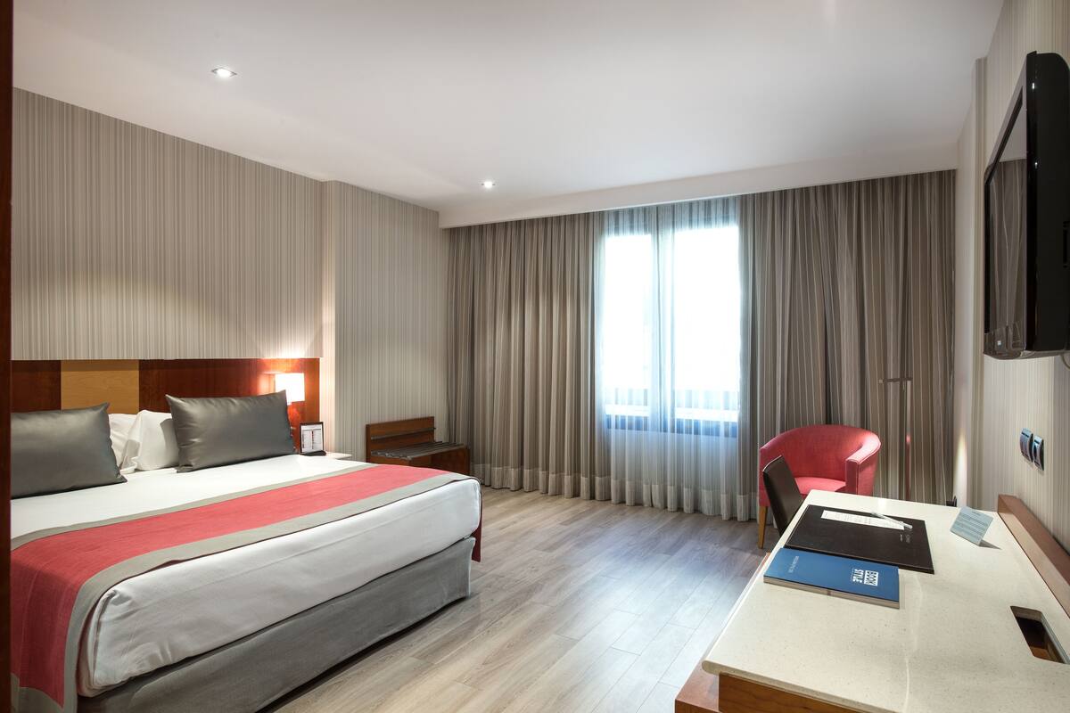 A hotel room with a large bed, a desk with a chair, a TV, and a window with curtains.