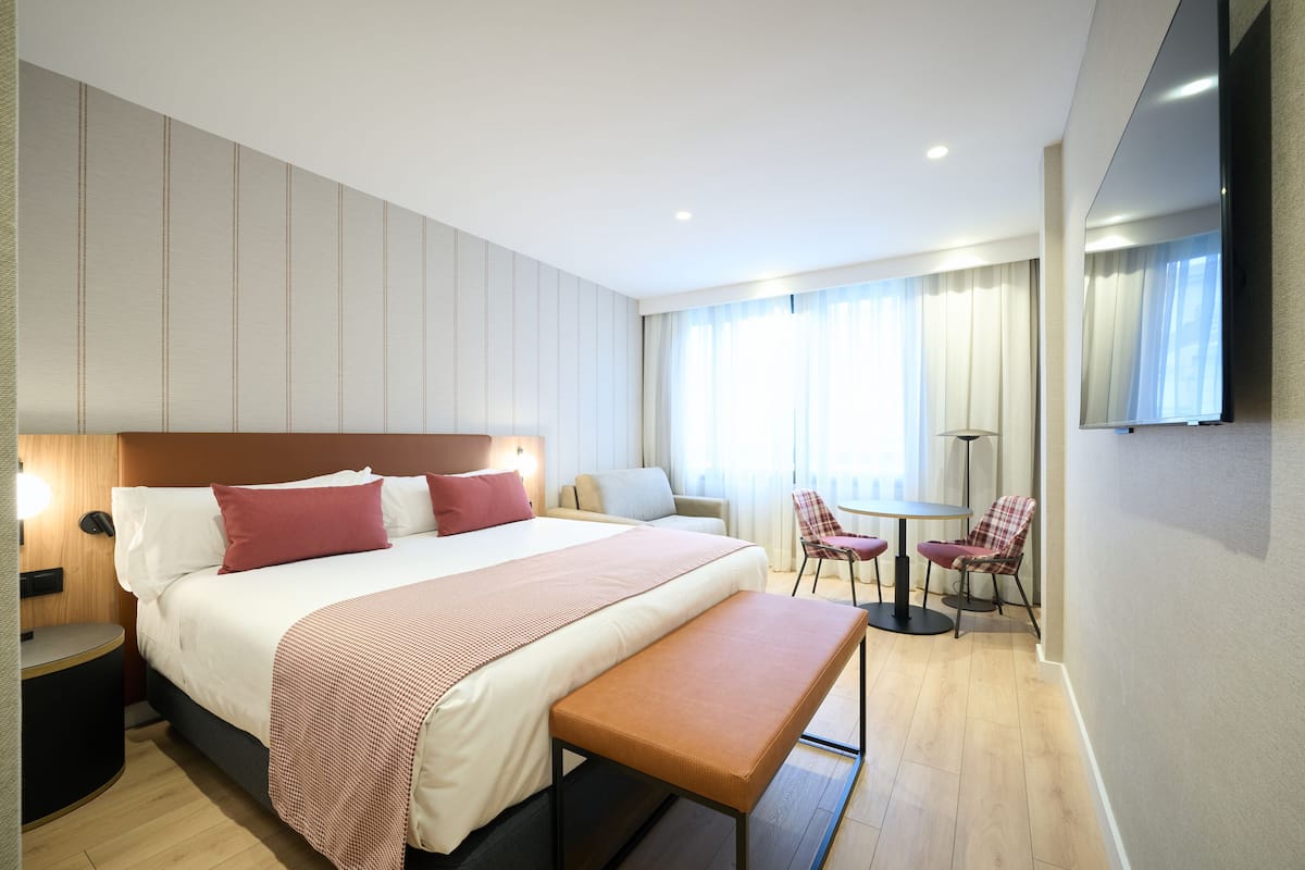 A modern hotel room with a large bed, bedside tables, a bench, a desk, and a chair.