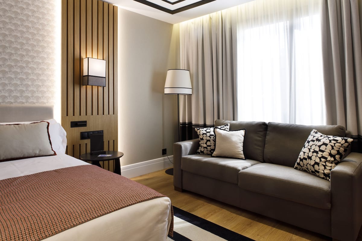 A hotel room with a bed, a sofa, a lamp, and a window with curtains.