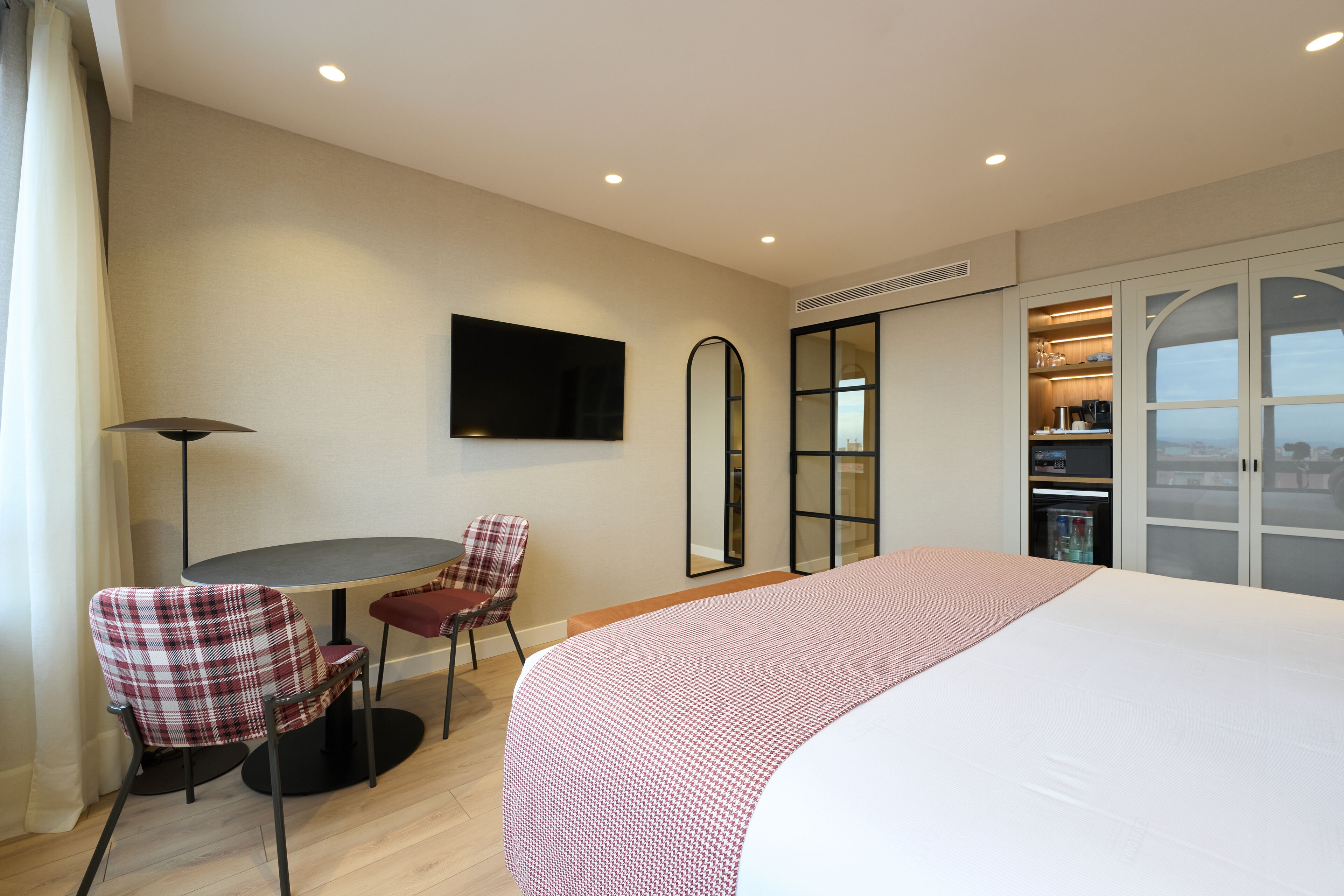 A hotel room with a large bed, a dining table with chairs, a TV mounted on the wall, and a wardrobe with a sliding door.