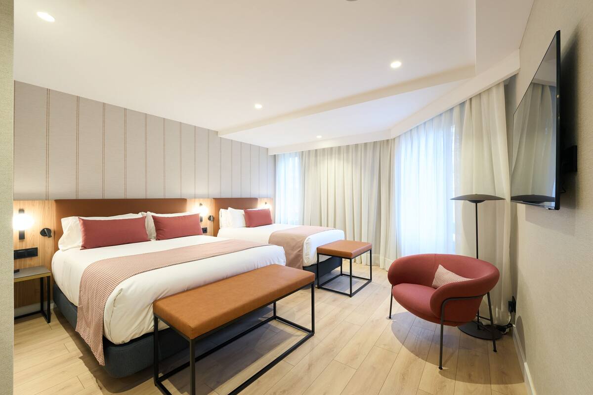A modern hotel room with two beds, a bench, a red chair, a TV, and a floor lamp.