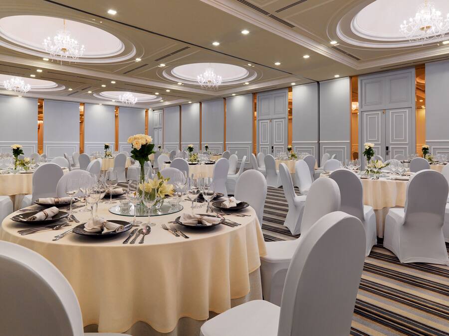 A banquet hall with round tables, white chairs, and chandeliers.
